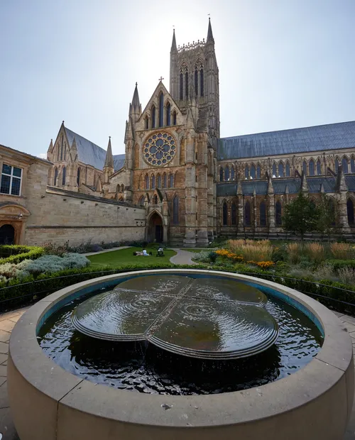 This architectural photograph, taken in the late morning during early autumn, features the water feature and fountain located in the grounds of Lincoln Cathedral in Lincoln, England, United Kingdom. The image prominently displays the historic cathedral, known for its Gothic architecture, with its large west front and iconic central tower rising above the scene. In the foreground, the circular fountain adds an element of stillness to the composition, its surface gently rippling beneath the sunlight. The cathedral, built as a place of Christian worship and an iconic church in England, is a well-known landmark with intricate stonework and the large rose window visible above the main entrance. The garden surrounding the fountain is in late summer bloom, contributing to the seasonal atmosphere of the photograph. The overall scene exemplifies English ecclesiastical architecture, connecting the fountain and landscaping with the historic structure of Lincoln Cathedral in an urban landscape setting.